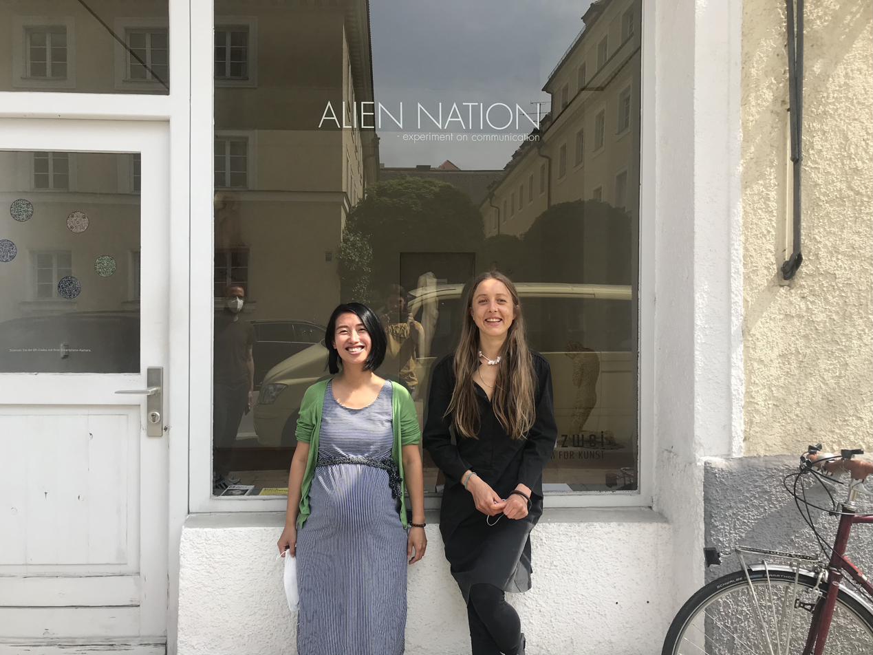 Two women lean smiling against a gallery window lettered Alien Nation, one in a green cardigan, the other in black