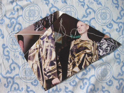 A woman with a shaved head in a gold patterned coat, green beads at her throat, folded to a point