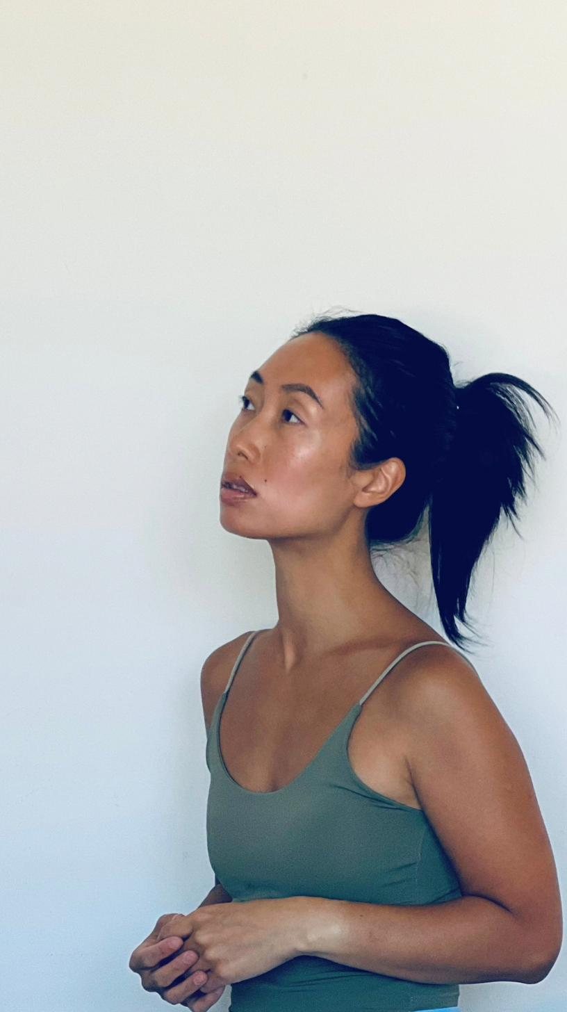 April Kam in profile against a pale wall, hair tied back, looking up and away from the camera